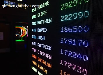 A digital leaderboard displaying names and scores in vibrant colors like green, blue, purple, and orange. The display is in a dark setting, possibly an arcade, and features a bright LED design.