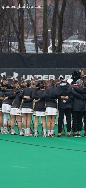A group of athletes is gathered in a huddle on a sports field, wearing matching uniforms and gear. The ground is green, and there is signage with a brand logo in the background. Trees and parked cars can be seen behind a fence surrounding the field. A person on the left side is capturing the moment with a camera.