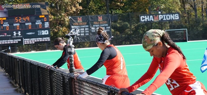A group of athletes wearing orange uniforms are on the sideline of a sports field, resting against a fence. Behind them, a large digital scoreboard displays team names and scores. The field is bright green, and several banners with sponsor logos and championship wins are visible. The athletes appear to be taking a break, possibly before or during a field hockey game.