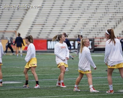 A group of young women wearing athletic gear are on a sports field. They are dressed in white jerseys with yellow shorts, featuring a distinctive design at the hem. The field has a green turf surface and the background shows empty bleachers. Some additional figures are visible in the distance, possibly referees or other players.