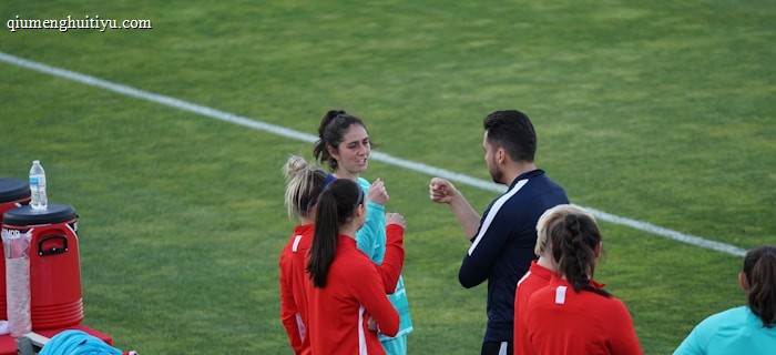A group of people wearing sports attire are gathered on a grassy field. Some individuals are dressed in red jackets, while others are in turquoise and navy blue. A coach appears to be giving instructions. Sports equipment, such as water bottles and a bench with towels, can be seen nearby.