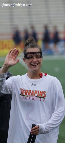 Several athletes in sports attire are on an outdoor sports field, interacting and smiling. One individual in the foreground waves with a friendly expression, while another stands beside her, also smiling. They wear protective goggles and gear associated with lacrosse. The background features indistinct figures in dark clothing against a backdrop of stadium seating.